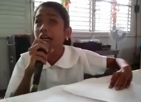 A young person is sitting in front of a table, holding a microphone in one hand, appearing to sing or speak. They are wearing a white shirt. The background shows windows with horizontal blinds, and some electronic equipment, like a fan and a speaker, is visible in the room. The atmosphere appears to be a casual or school setting.