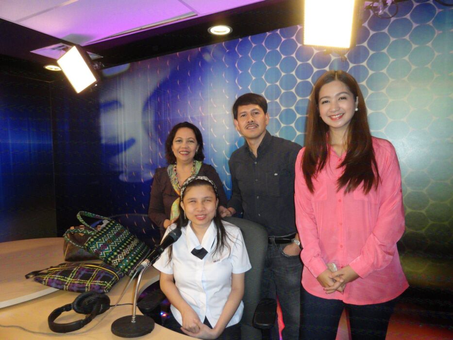 Four people in a radio studio. In the foreground, a young woman in a white uniform sits at a table with a microphone. Behind her, three people stand: a woman with short hair wearing a patterned scarf and dark clothing, a man with facial hair in a blue shirt, and a smiling woman with long hair wearing a pink blouse. The studio is well-lit, featuring walls with a blue honeycomb pattern. A pair of headphones is also visible on the table.