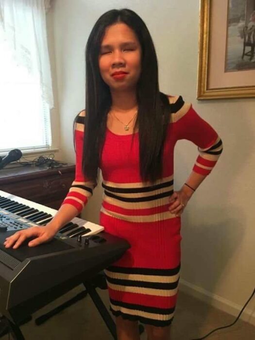 A woman with long black hair wearing a red dress with horizontal white and black stripes. She is standing beside an electronic keyboard, and her right hand is resting on it. The room appears to be a living room, with a framed picture on the wall and a window with white curtains in the background.