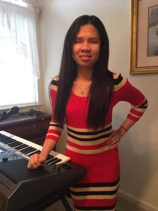 A woman with long dark hair standing next to an electronic keyboard. She is wearing a fitted red dress with black and cream horizontal stripes. The room has light-colored walls, a window with white curtains, and a framed picture hanging on the wall. The woman is smiling slightly and has one hand resting on the keyboard and the other hand on her hip. The setting appears to be indoors, possibly a living room or music room.