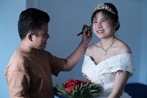 A joyful bride in an off-the-shoulder white wedding dress with a floral pattern. She is wearing a golden tiara, a matching necklace, and earrings. She is holding a bouquet of red roses. A person in a tan shirt is applying makeup to her face with a brush, causing her to smile widely. The background appears to be a simple, light blue setting, which emphasizes the central interaction.