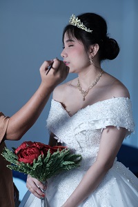 The image shows a bride in a white, off-the-shoulder wedding gown, holding a bouquet of red flowers with green leaves. She is wearing a gold crown and matching jewelry. A person is applying makeup to her face, focusing on her lips. The background is plain and blue. The bride appears calm and poised.